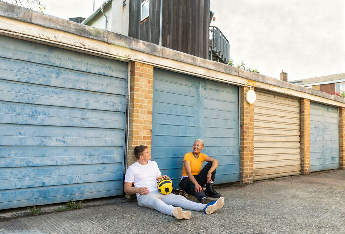 Two adults sitting on the ground outside garages, smiling and holding a yellow and black volleyball, childhood memories.
