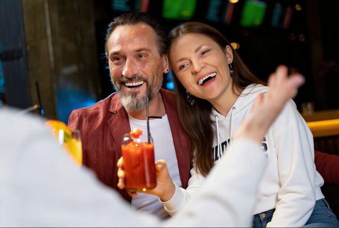 A man with a beard and a woman laughing joyfully at a bar, sharing wildest rumors over drinks.