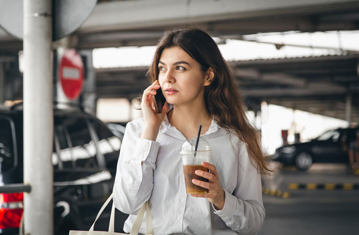 Young woman holding iced coffee, talking on phone in parking garage, relating to single mom and autistic kid caregiving challenges.
