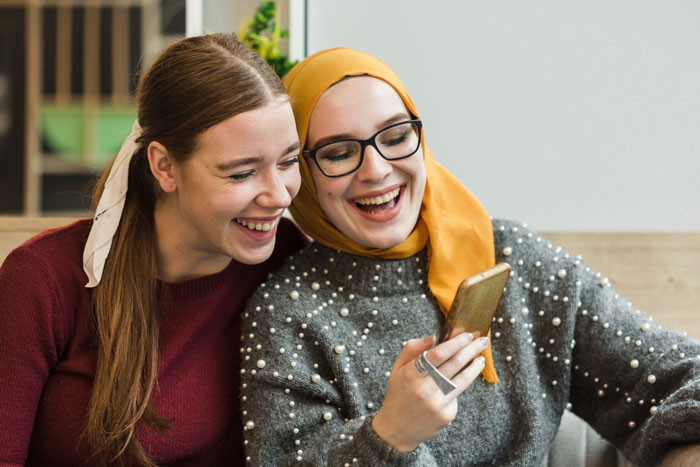 Two smiling friends, a Christian bride and a Muslim best friend, share a laugh while looking at a phone.