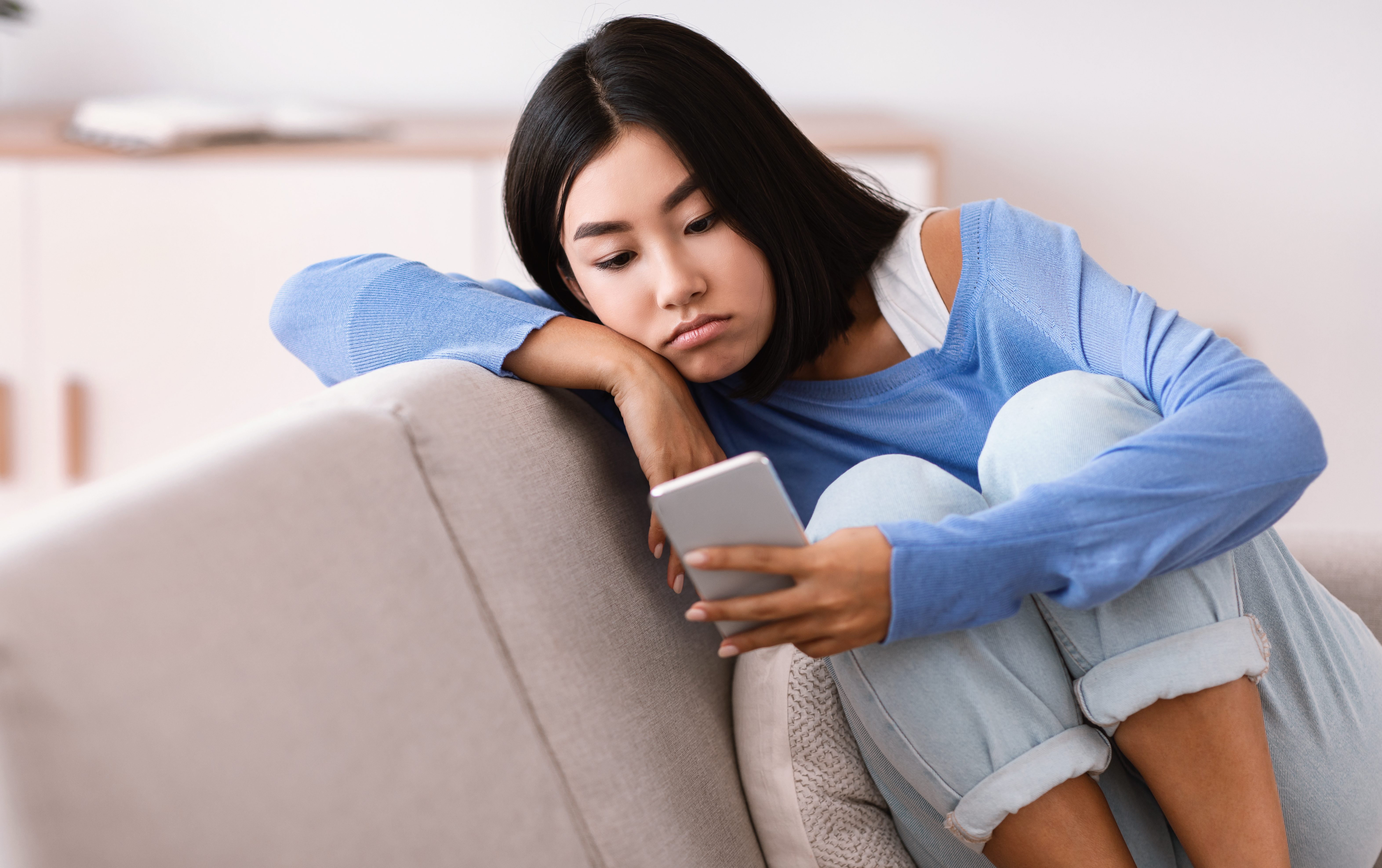 A sad woman on a couch, holding a phone, upset her best friend skips wedding due to not feeling well.
