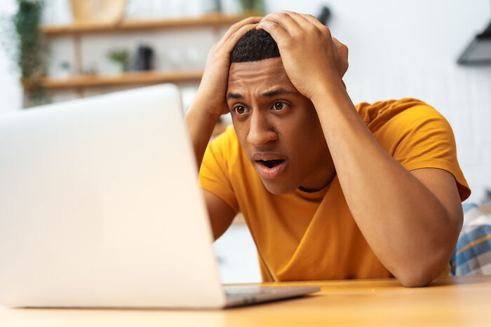 A man in a yellow shirt looks shocked at his laptop, hands on his head, after finding out he was being cheated on.