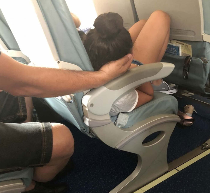 Woman sleeping with legs curled up on airplane seat while a man’s hand supports her head in a funny sleeping position.
