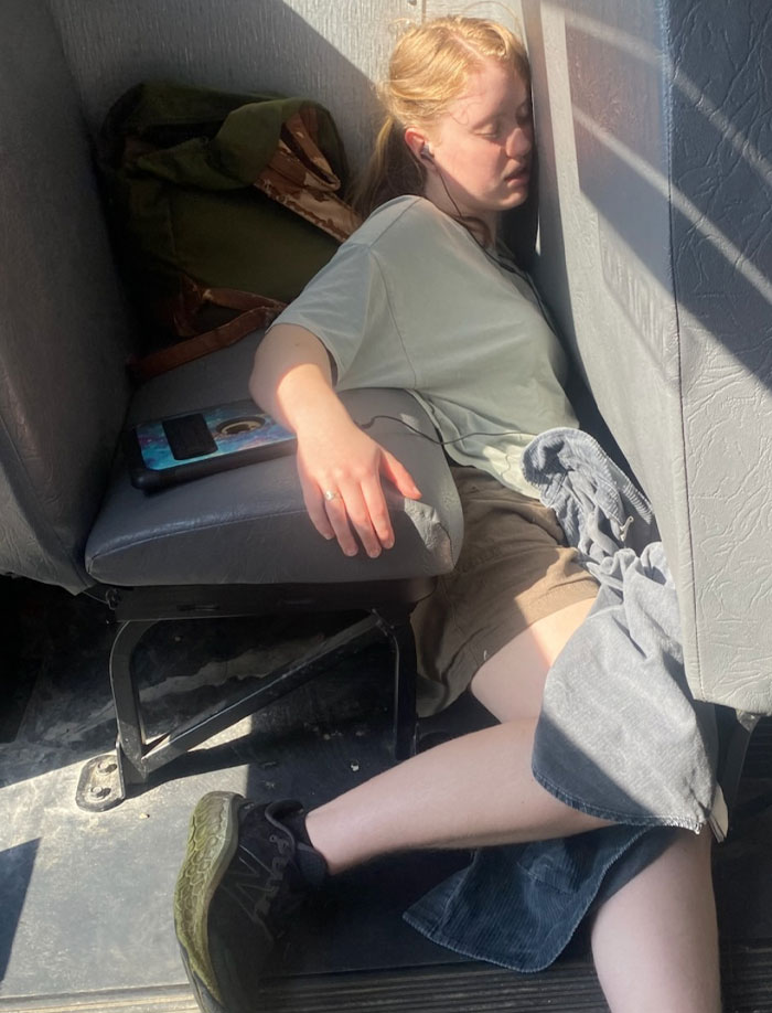 Person caught sleeping unhinged on a bus floor, leaning against seats with a phone nearby in a bright sunlit setting.
