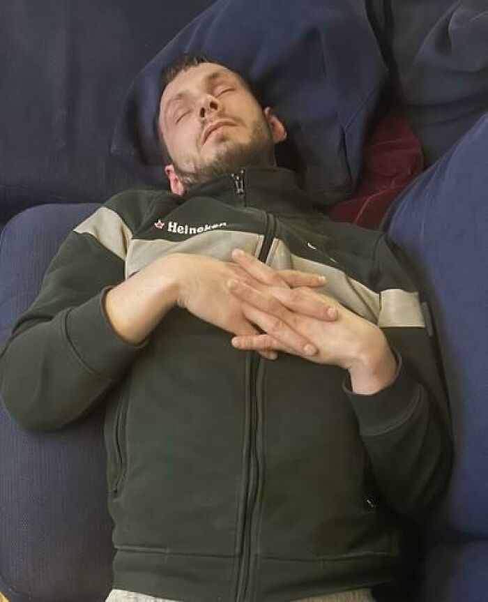Man caught sleeping in a hilarious and unhinged way, lying with hands folded on chest on a couch.