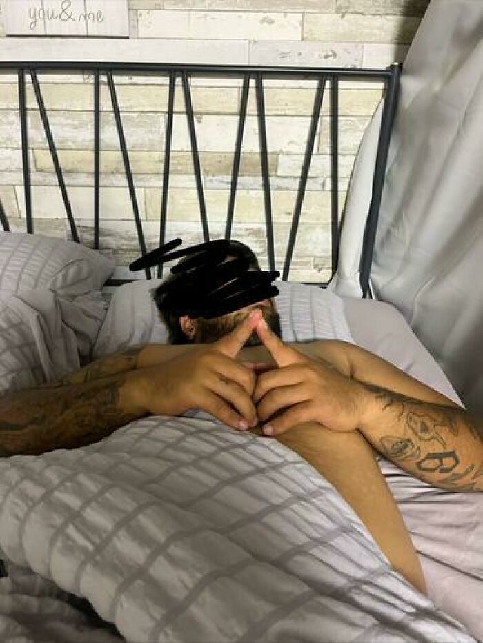 Person caught sleeping in a bed with tattoos on arms, lying under white bedding in a relaxed and unhinged pose.