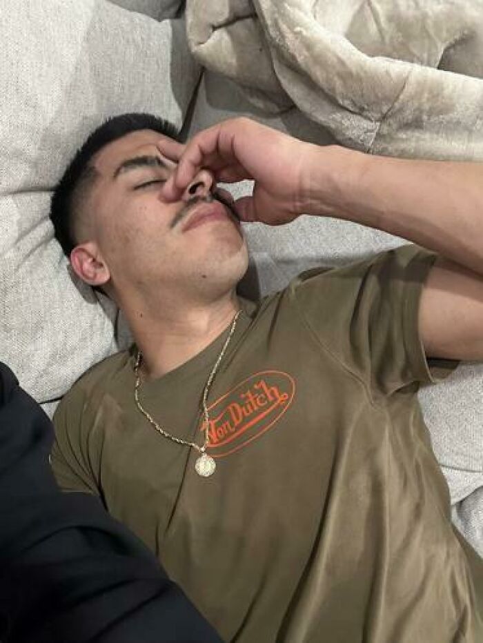 Young man caught sleeping in a hilarious way on a couch, wearing a green Von Dutch shirt and a gold chain necklace.
