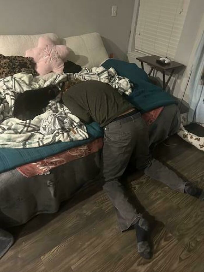 Person caught sleeping in an unhinged position with head on bed and knees on floor next to a black cat on the couch.