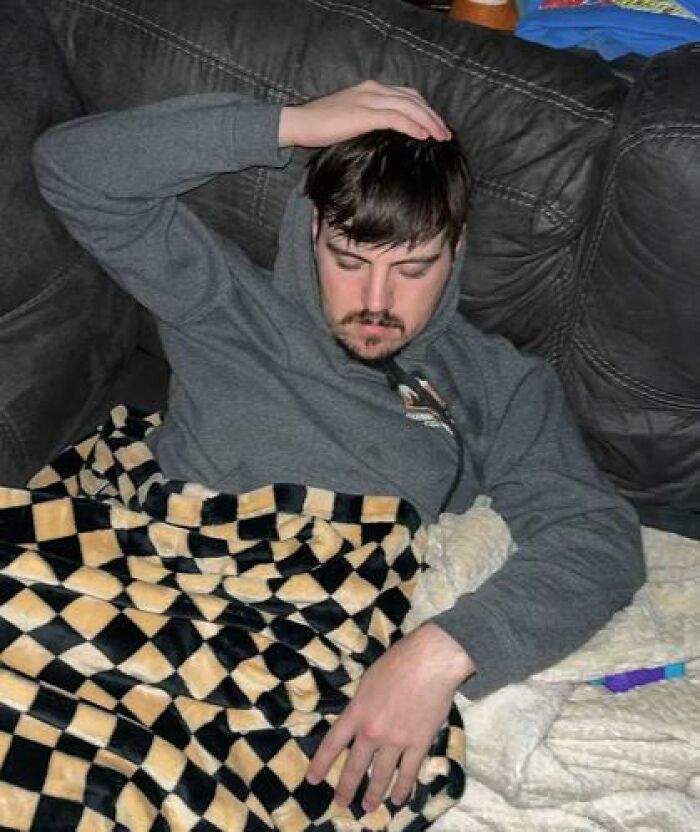 Man caught sleeping in hilarious way on couch with checkered blanket and gray hoodie resting hand on head.