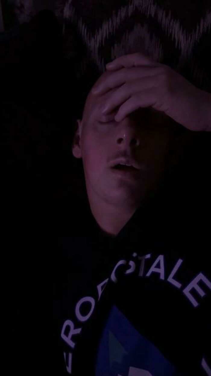Young man caught sleeping in a dark room with hand on forehead, one of the hilarious and unhinged sleeping moments captured.