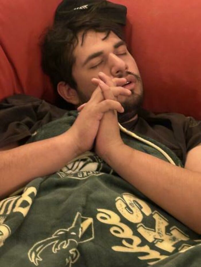 Young man caught sleeping in hilarious and unhinged ways, resting with hands clasped on a couch with a blanket.