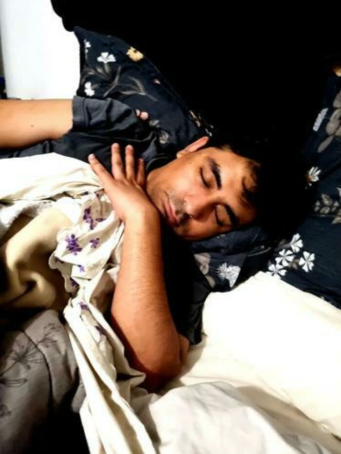 Man caught sleeping in hilarious and unhinged ways, lying on bed with floral pillow and blanket covering part of his body.