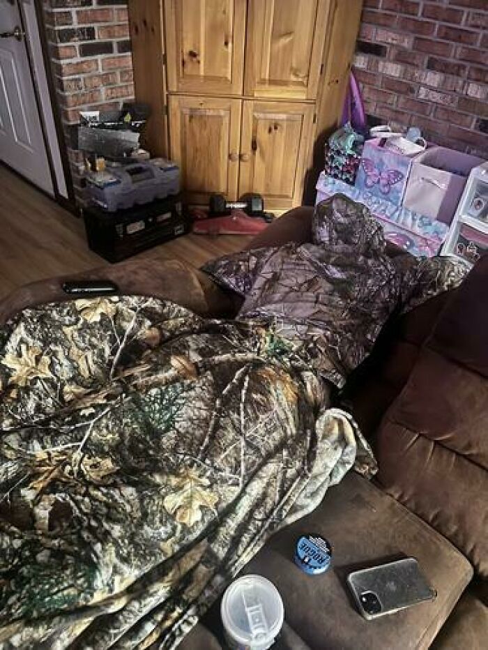 Person caught sleeping wrapped entirely in camouflage blanket and hood on a couch in an indoor setting.
