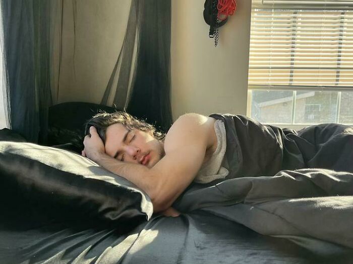 Young man caught sleeping in an unhinged way, lying on black bedding near a sunlit window in a bedroom.