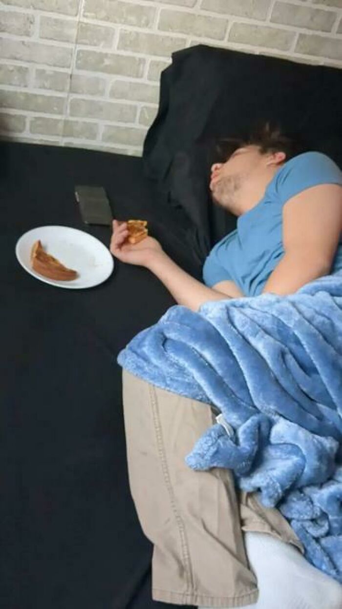 Person caught sleeping while holding a slice of pizza on a black bed with a plate and phone nearby under a blue blanket