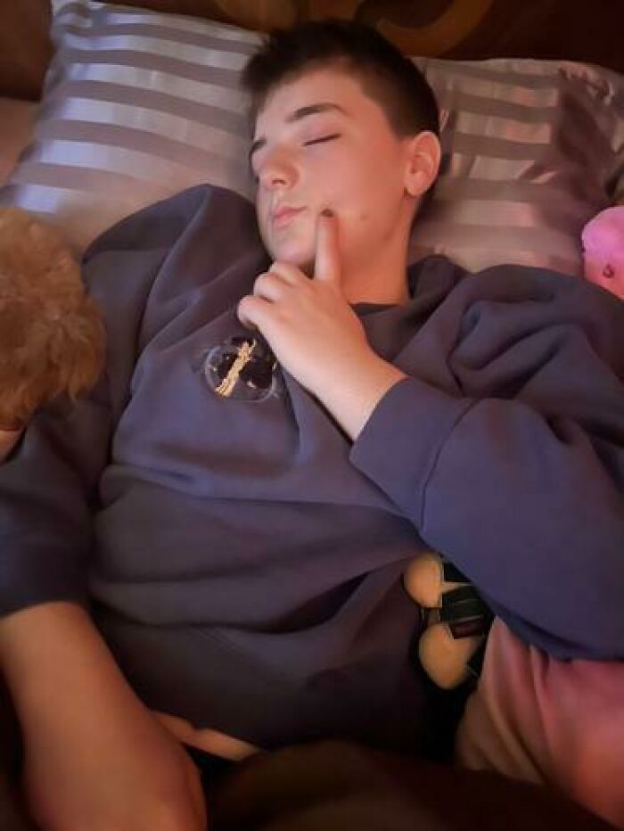 Teen caught sleeping in hilarious and unhinged way with finger resting on cheek, lying on striped pillow and holding a plush toy.