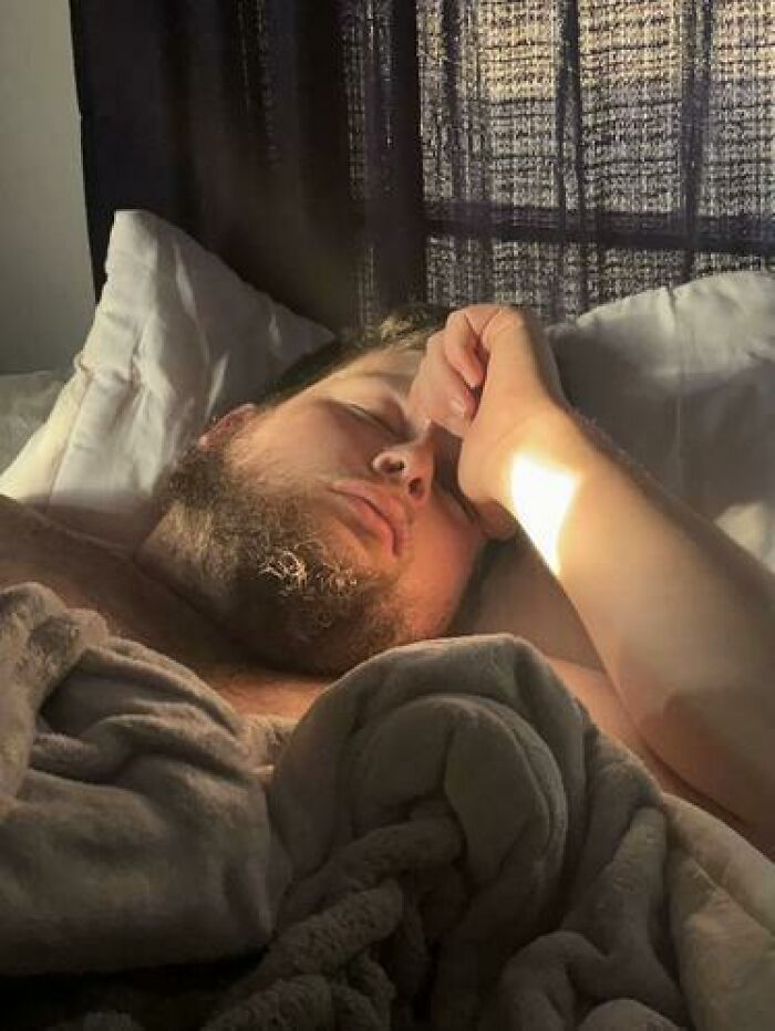 Man caught sleeping in a funny way, resting with arm over face under a blanket in soft morning light near a window.