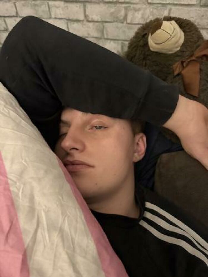 Young man caught sleeping in a funny position with arm over his face and a stuffed bear in the background.