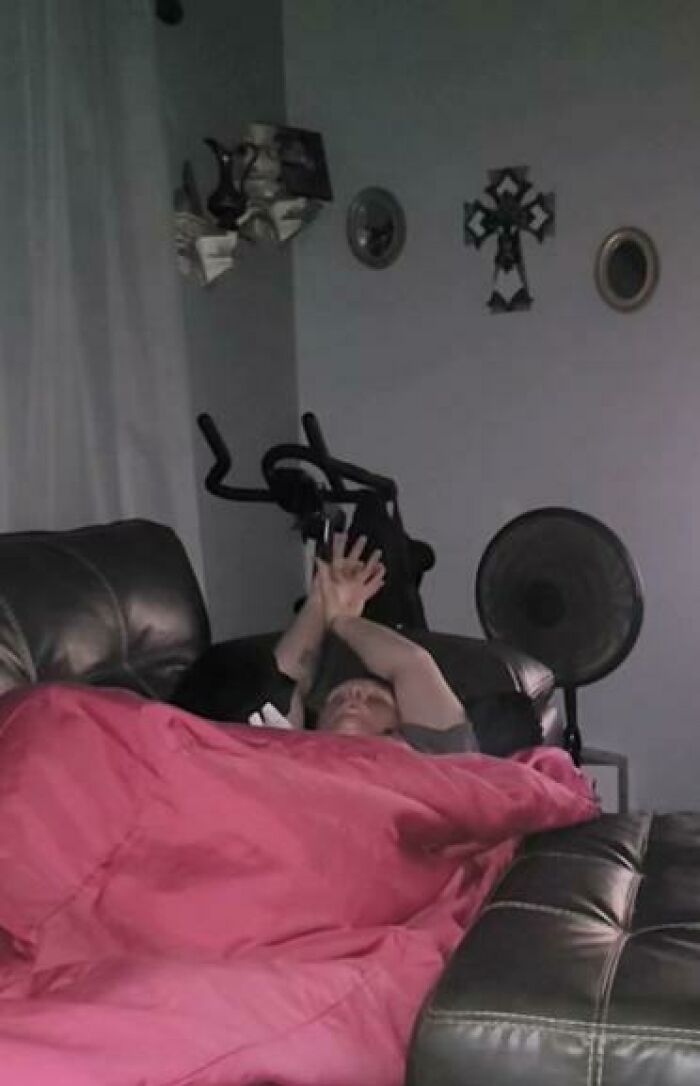 Person caught sleeping on a couch covered with a pink blanket, hands raised, next to a fan and exercise equipment indoors.