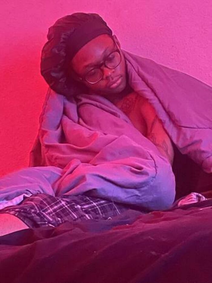 Person caught sleeping wrapped in a blanket and wearing a bonnet and glasses under pink lighting indoors.