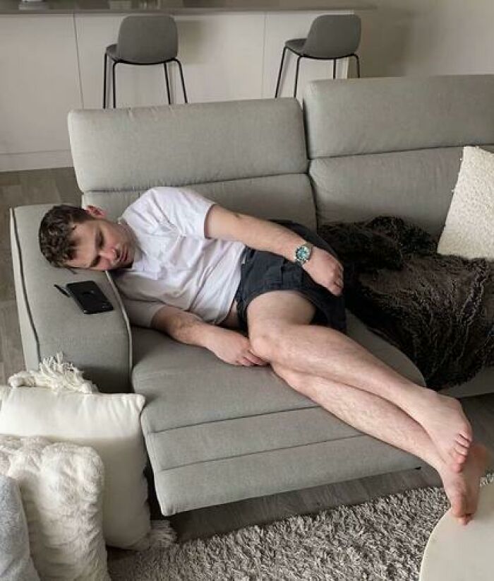 Man caught sleeping in hilarious and unhinged ways, lying sideways on a couch with phone nearby in a modern living room.