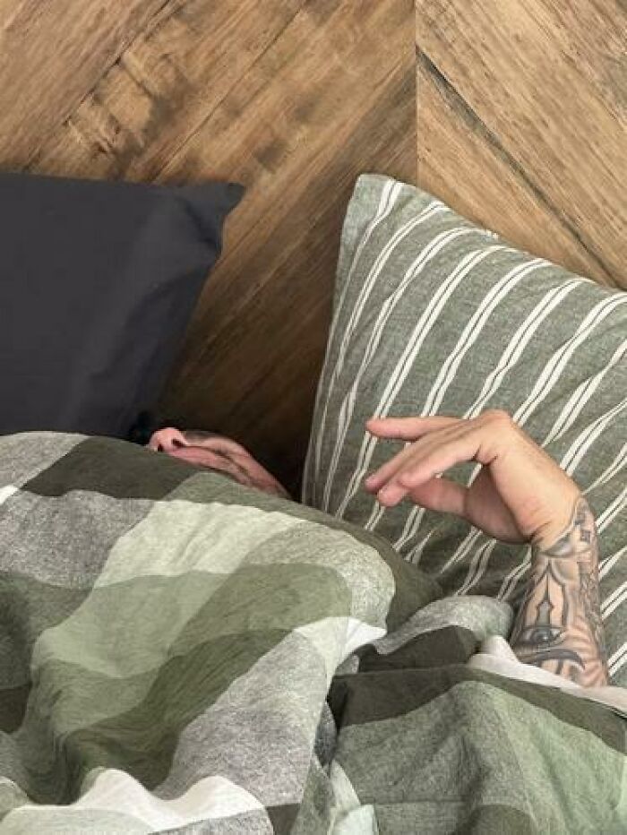 Person caught sleeping in a bed with patterned pillows and a green camouflage blanket, hand resting visibly with tattoos.