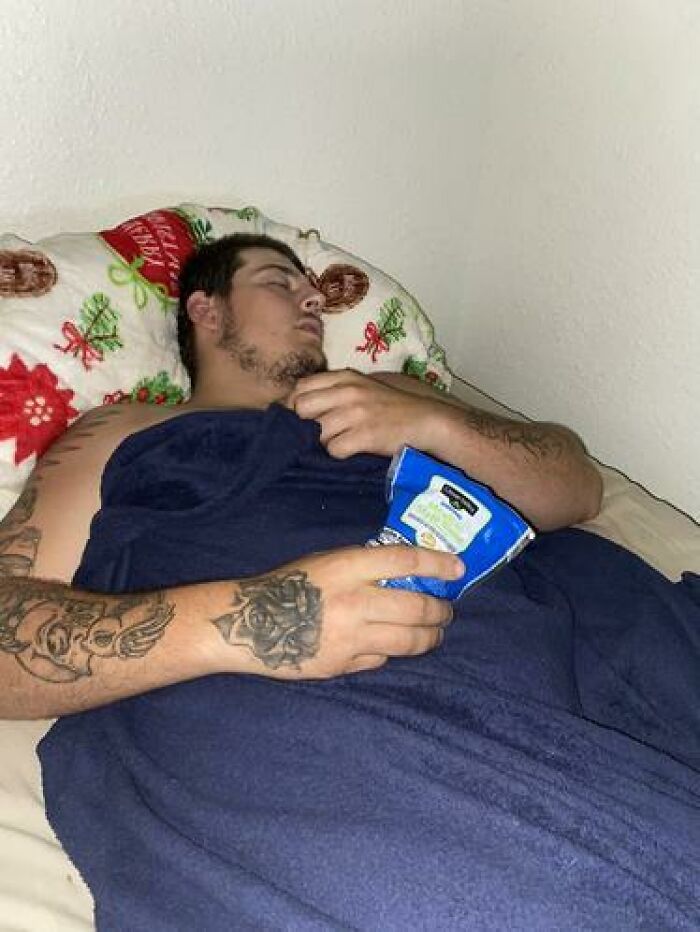 Man caught sleeping in hilarious and unhinged ways, holding a chip bag while lying on a festive pillow and covered with a blanket.