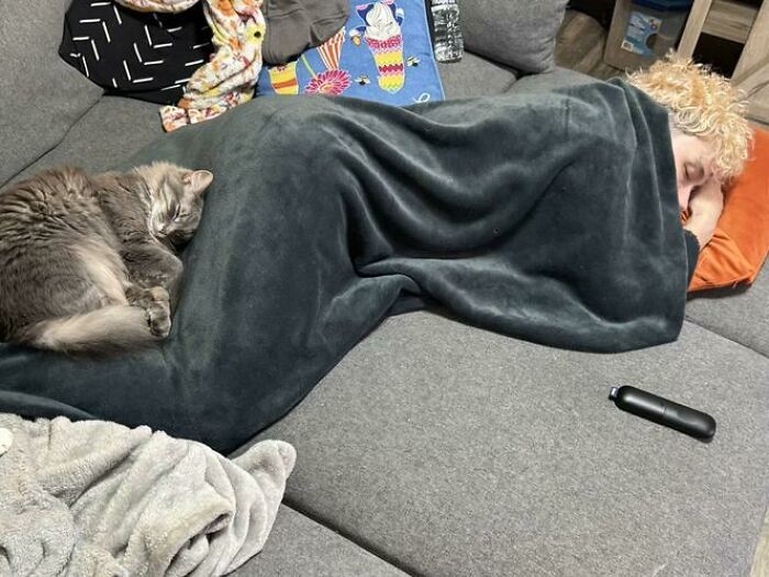 Person and cat caught sleeping in hilarious and unhinged ways on a couch covered with a dark blanket and colorful pillows.