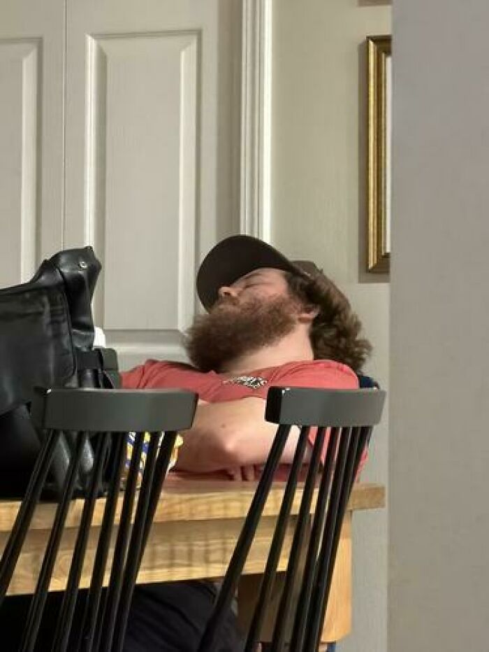 Man with a beard and cap caught sleeping in an unhinged way, slumped over a table behind black chairs indoors