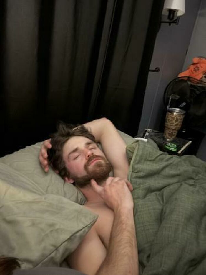 A man caught sleeping in a relaxed pose with arm behind head, covered by green bedding in a dimly lit room.