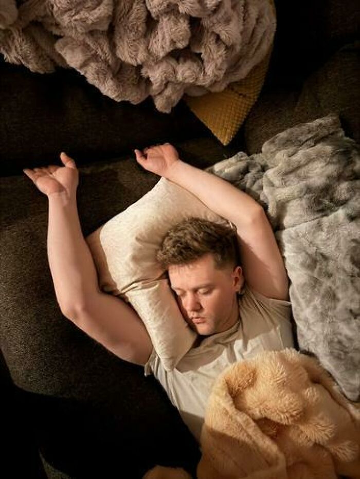 Young man caught sleeping in hilarious and unhinged ways with arms raised, surrounded by soft blankets and pillows on a couch.