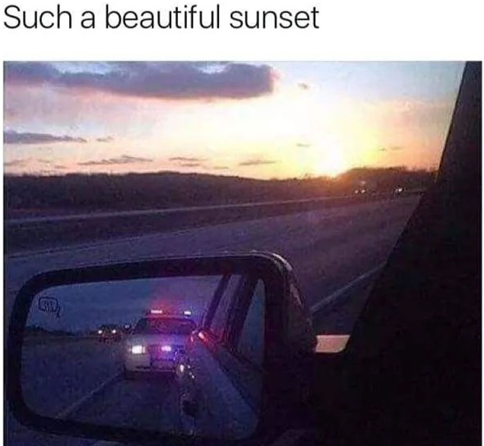 Rearview mirror showing police car with flashing lights behind during a sunset, a funny and lighthearted safe-for-work meme.