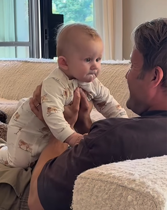 Gay dad holding surrogate baby on couch, sharing heartfelt moment amid backlash over video controversy.