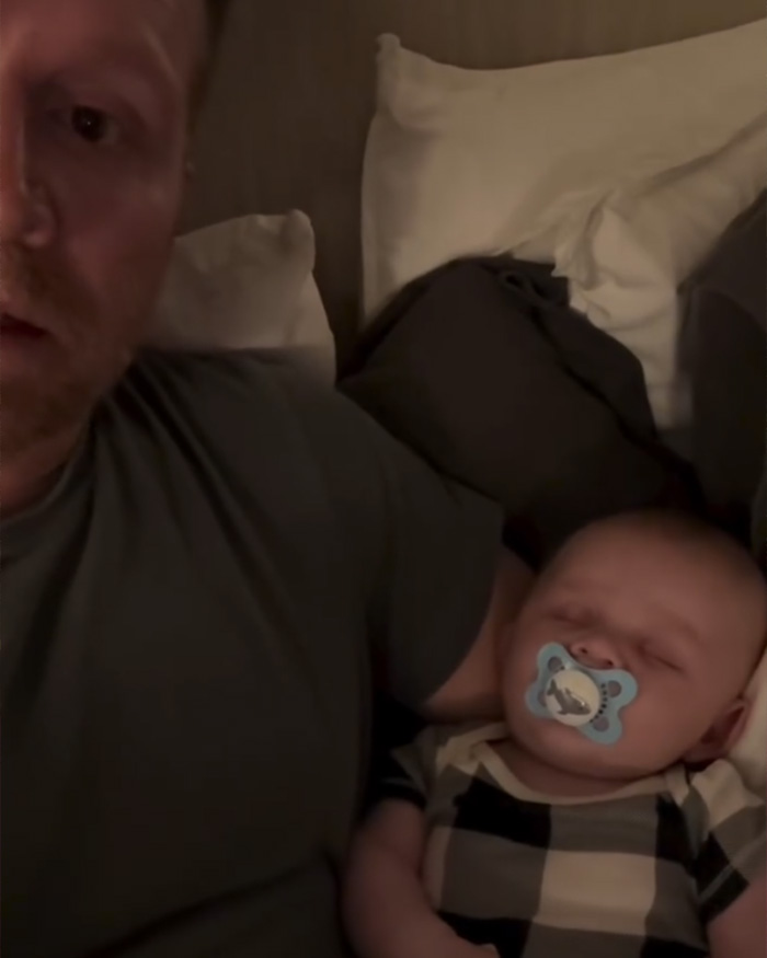 Gay dad sitting on a bed holding a sleeping baby with a pacifier amid backlash over surrogate baby video.