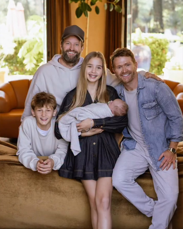 Gay dad and family posing together indoors, smiling with a baby wrapped in a blanket in the center.