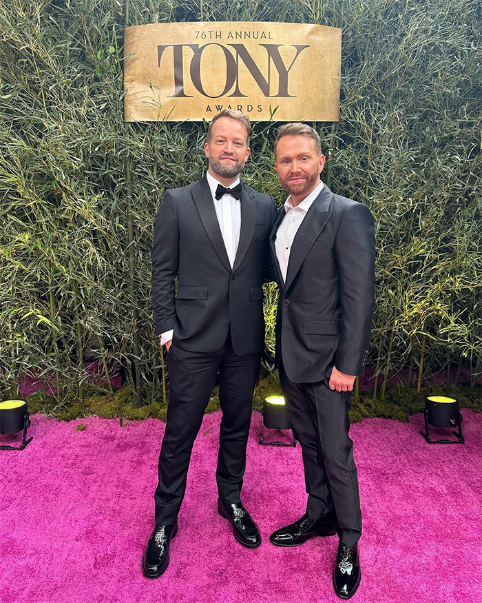 Two men dressed in black tuxedos posing on a pink carpet at the 76th Annual Tony Awards event.