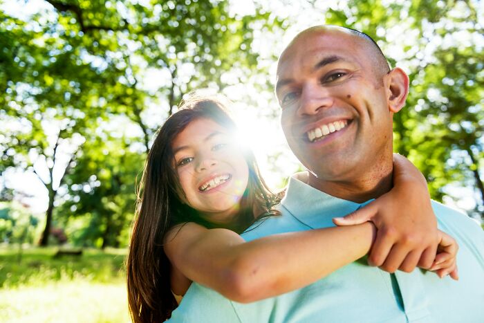 A happy dad and daughter smiling together outdoors, sunlight peeking through trees. Raising daughters.