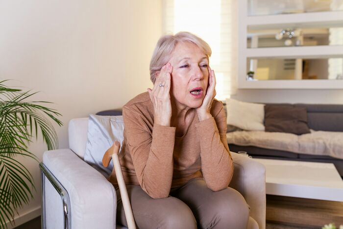 An elderly woman with blonde hair, wearing a brown shirt, looking distressed with hands to her temples, conveying a "worst MIL" story.