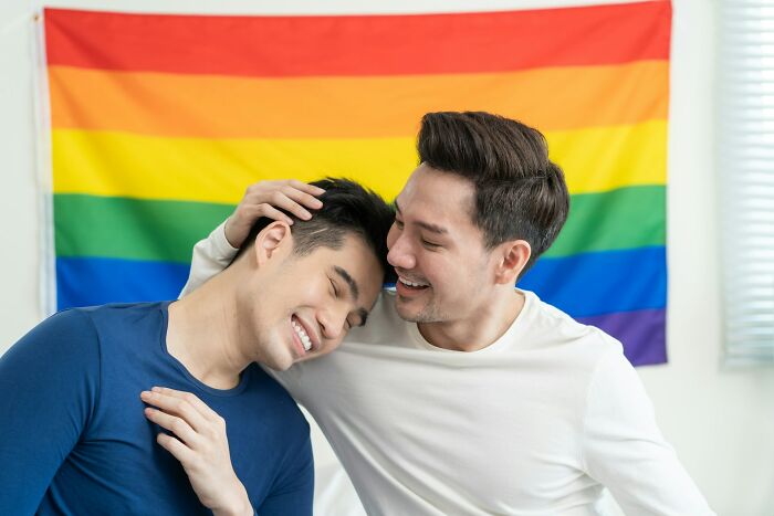 Two men sharing a tender moment in front of a rainbow flag, showing family love in unexpected ways.