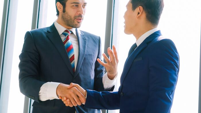 Two businessmen in suits shaking hands and discussing ideas, displaying intelligence in a professional setting.