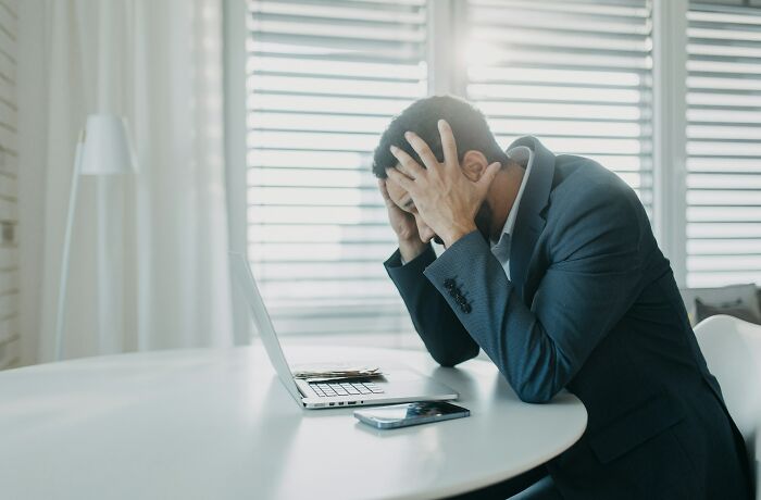 Man in suit holding his head in frustration after accidentally sending out a mortifying message on laptop at a desk