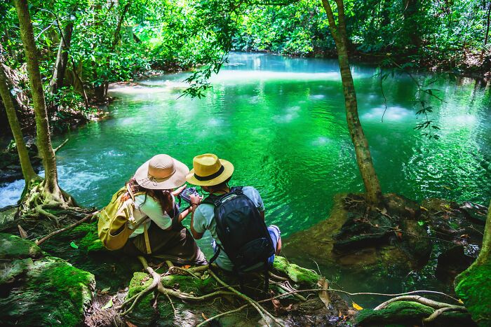 Two tourists with hats and backpacks near a vibrant green lake in a lush forest, enjoying their time abroad.