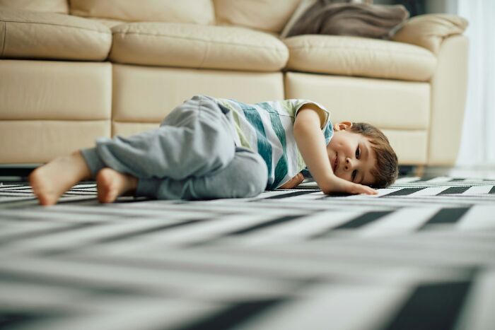 Young boy lying on a patterned rug in the living room, reflecting on normal family activities that seem bizarre now.