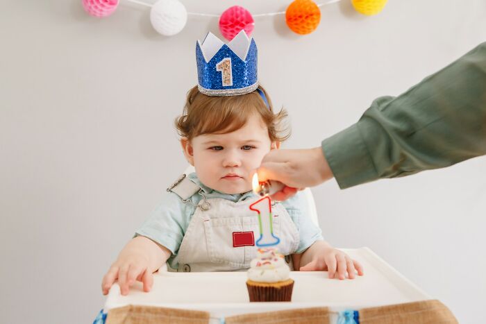 A baby with a sparkly blue crown looking unenthusiastic as a hand lights a 1st birthday candle on a cupcake. Worst MIL.