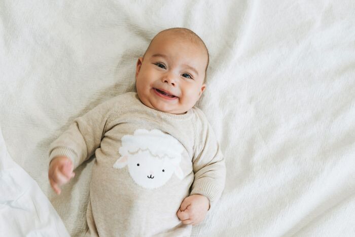 A smiling baby in a sheep-themed onesie, lying on a white blanket. Features "Worst MIL" stories.