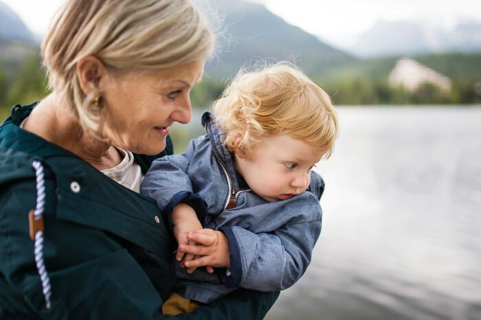 A woman holding a baby outdoors with a lake and mountains in the background. MIL stories come to mind.