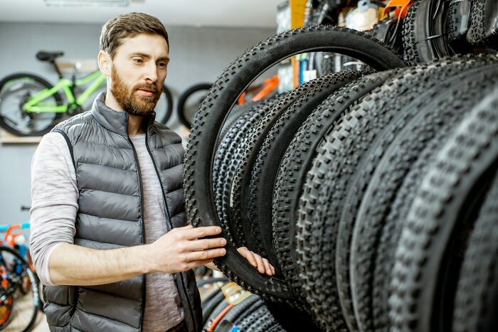Man in a gray vest examining bike tires in a store, surrounded by mountain bike wheels on display racks.