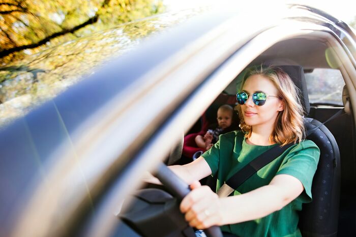 Woman wearing sunglasses driving a car with a child in the backseat, illustrating parenting tips and tricks shared by parents.