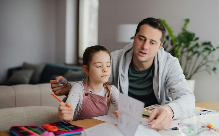 A dad helping his daughter with homework, both focused on a book and colored pencils. Raising daughters.