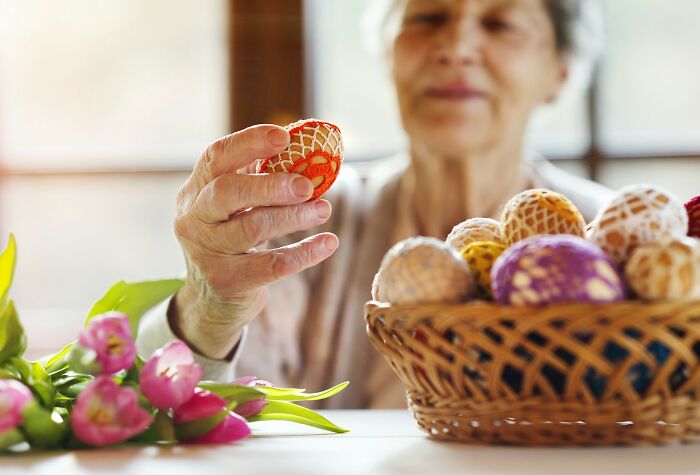 An elderly woman with gray hair holds a decorative, crocheted egg, with a basket of similar colorful eggs and pink tulips nearby. Worst MIL stories.
